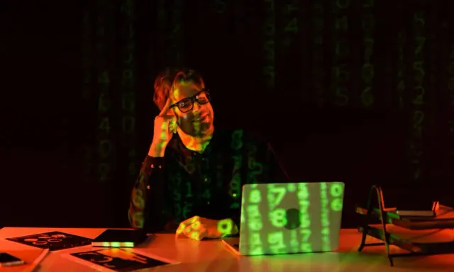 Mężczyzna w okularach siedzi przy biurku, na którym leży laptop z zielonym kodem.