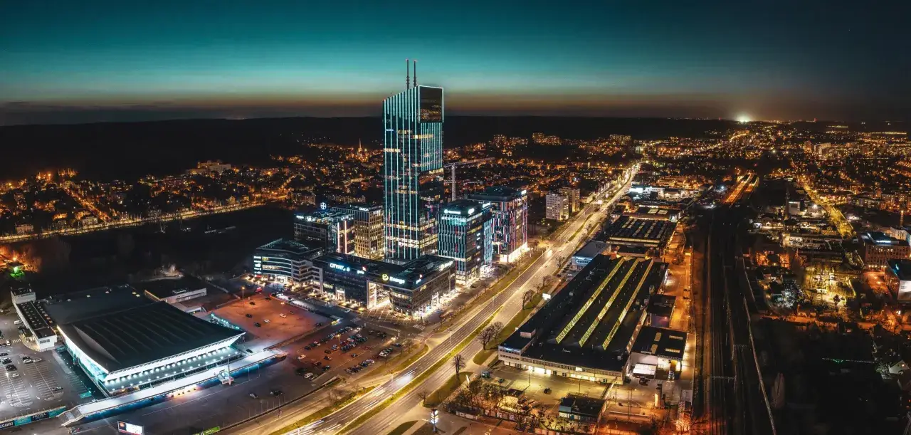 Widok z lotu ptaka na nowoczesne centrum miasta nocą, z oświetlonymi wieżowcami i ulicami.